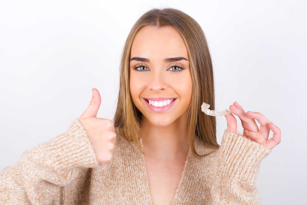 Kobieta z białymi zębami pokazuje kciuk w górę i trzyma Invisalign w ręku